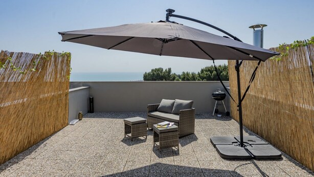 Una terrazza sul tetto con un divano in vimini, due tavolini, un ombrellone grande e un barbecue.