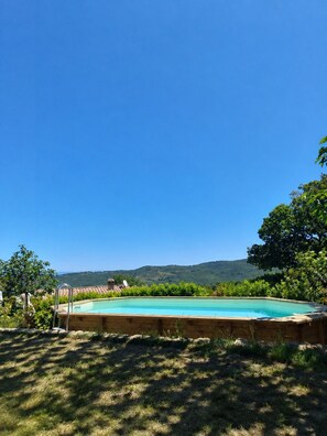 Ein Schwimmbecken mit Holzterrasse, umgeben von Gras und Bäumen, mit blauem Himmel und Hügeln im Hintergrund.