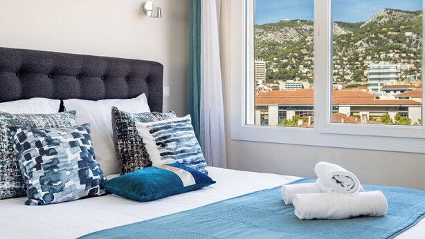 Habitación de hotel con cama, almohadas, ventana y vistas a un paisaje urbano y montañas.