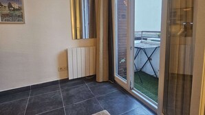 Une pièce dotée d’un sol carrelé, d’un radiateur, d’une porte-fenêtre coulissante donnant sur un espace extérieur avec une table et des chaises, et d’un tableau encadré accroché au mur.