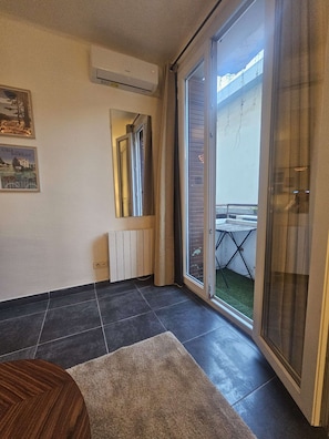 Ein Zimmer mit Fliesenboden, einem Heizkörper, einer Schiebetür zu einem Außenbereich mit Tisch und Stühlen sowie einem gerahmten Bild an der Wand.