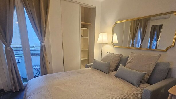 Une chambre à coucher avec un grand lit, une lampe de chevet, un miroir et une vue sur l’extérieur par une fenêtre avec des rideaux.