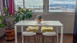 Un coin repas avec une table blanche et deux chaises, une plante en pot et une vue sur un port avec des grues.