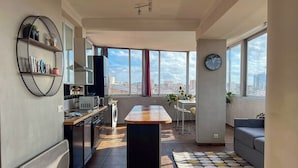 Une cuisine moderne dotée d’un îlot central, un coin repas avec une table et des chaises, un canapé et de grandes fenêtres offrant une vue sur la ville.