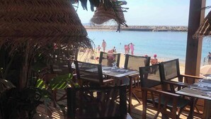 An outdoor dining area with tables and chairs, overlooking a beach with people and a clear sky.