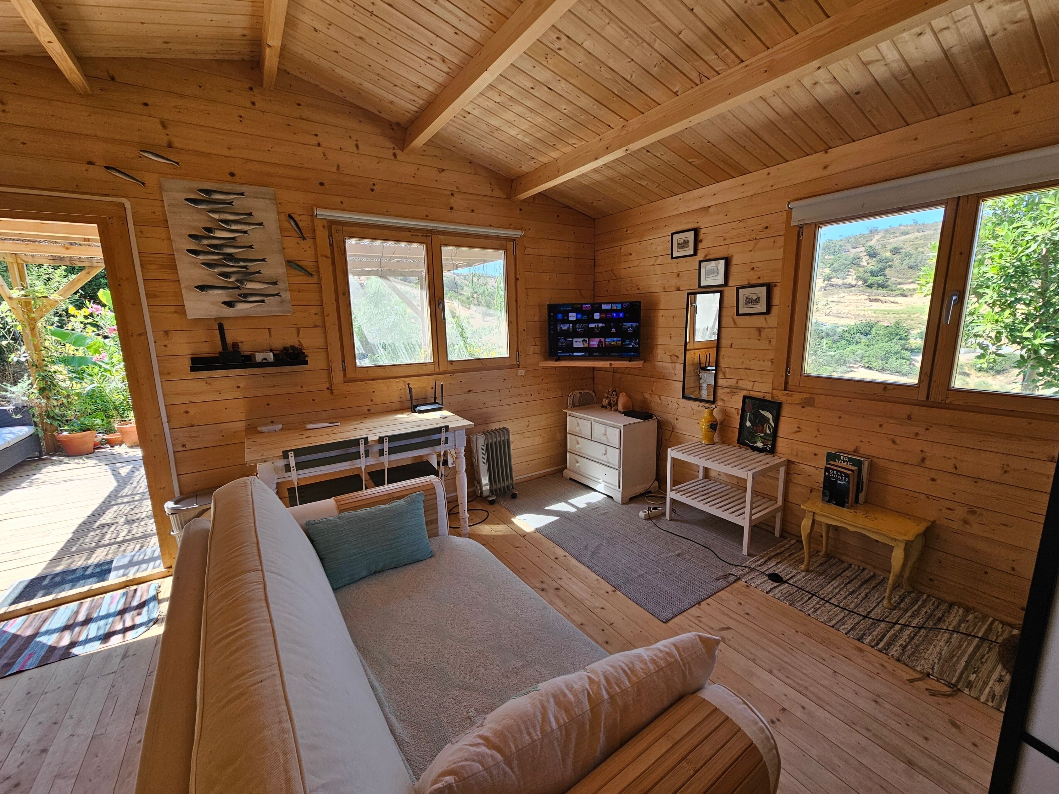 A wooden cabin interior with a sofa, a small table, a TV, and a window overlooking greenery.