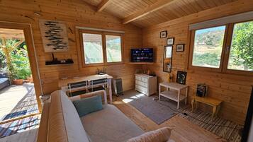 A wooden cabin interior with a sofa, a small table, a TV, and a window overlooking greenery.