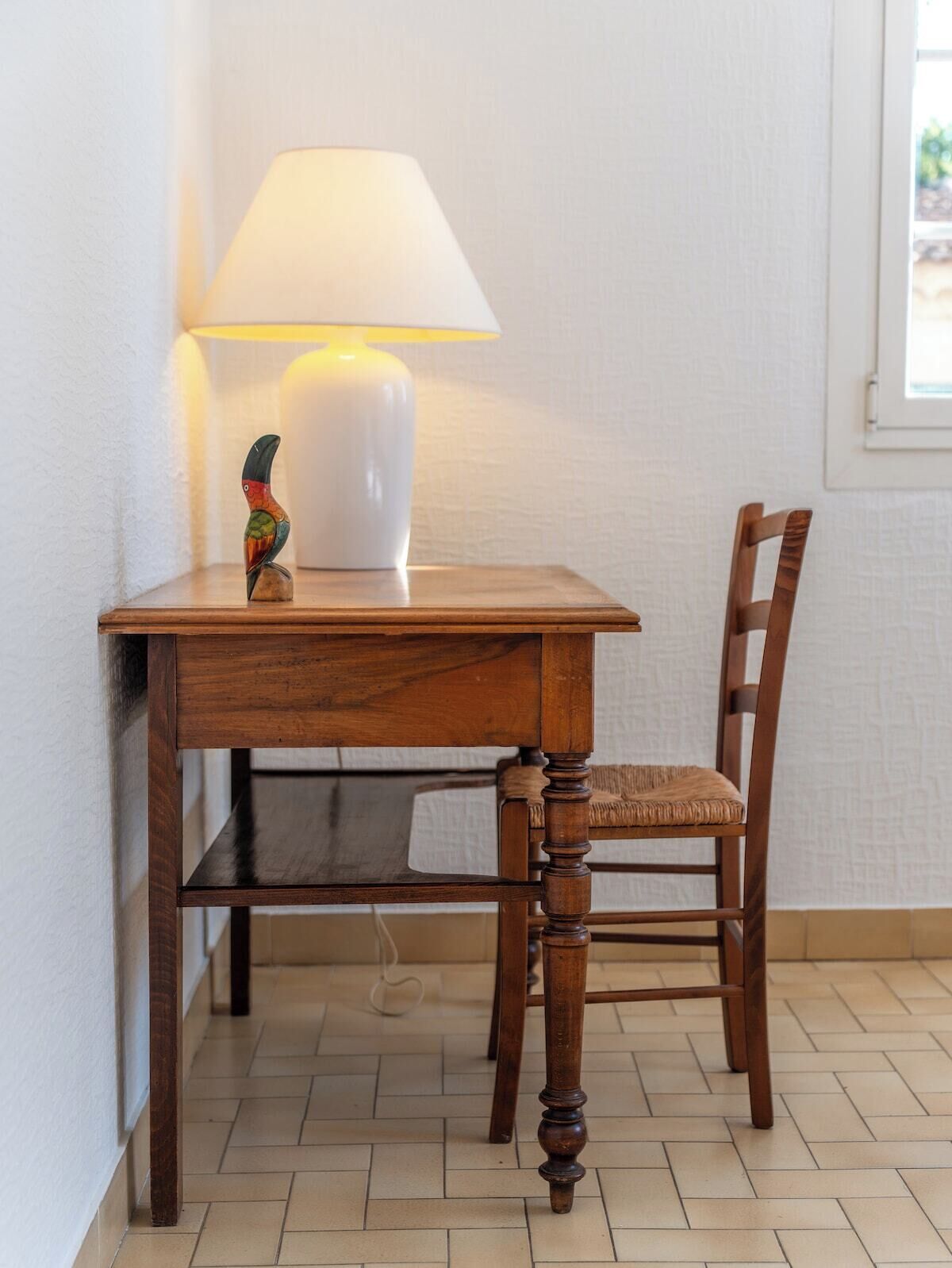 Een houten tafel met een lamp en een stoel in een kamer met betegelde vloer en een raam.