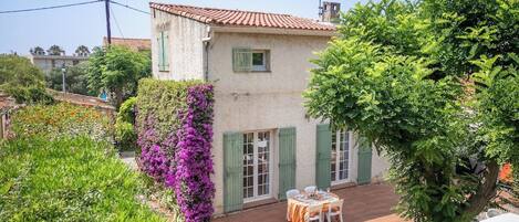 A two-story building with a tiled roof, green shutters, and a patio with a table and chairs.