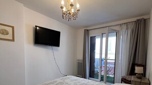 A bedroom with a bed, a television, a chandelier, and a view of the outdoors through a sliding door. - Joli T3 spacieux & climatisé – confort + wifi (Toulon)