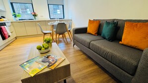 En moderne stue med en grå sofa, et træbord med blade, en skål med æbler og en lille plante.