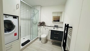 Et lille badeværelse med et glasafskærmet brusebad, toilet, vask med spejl og en vaskemaskine.