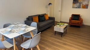En moderne stue med spiseområde, grå sofa med orange puder, et lille træsofabord og en enkelt orange lænestol.