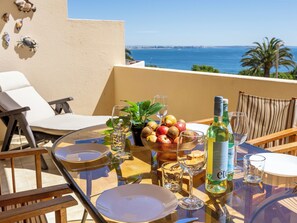 Ein Tisch mit Tellern, Gläsern, einer Flasche und Früchten auf einem Balkon mit Blick aufs Meer.