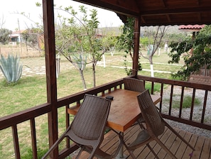 Überdachte Terrasse mit Tisch und Stühlen, mit Blick auf einen Garten mit verschiedenen Pflanzen und ein entferntes Gebäude.