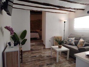 Sala de estar com sofá, uma mesinha, planta em vaso e uma cama ao fundo.