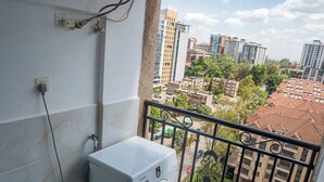 Ein kleiner Balkon mit Waschmaschine, blauem Eimer und Blick auf eine Stadtlandschaft.