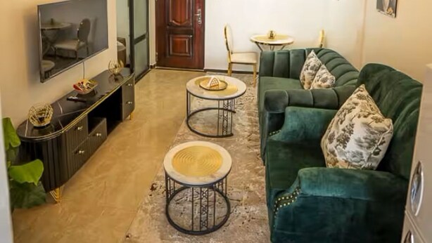 Ein Wohnzimmer mit einem grünen Sofa, einem Fernseher, einer Chandelüster und einem Essbereich.