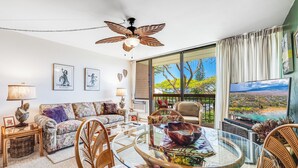 Un salon avec une table en verre, des fauteuils en osier, un ventilateur de plafond et une grande fenêtre donnant sur un beau paysage.