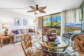 A living room with a glass table, wicker chairs, a ceiling fan, and a large window with a view. - Maui Vista #2-218 Close to Beach, Pickleball Courts, Full A/C (Kihei)