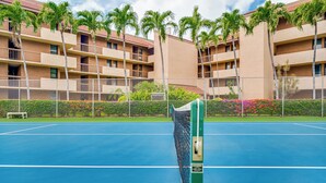 Un court de tennis avec un filet, entouré d’une clôture, et un bâtiment avec des balcons et des palmiers en arrière-plan.