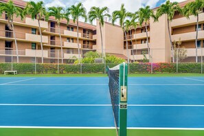 A tennis court with a net, surrounded by a fence, and a building with balconies and palm trees in the background. - Maui Vista #2-218 Close to Beach, Pickleball Courts, Full A/C (Kihei)