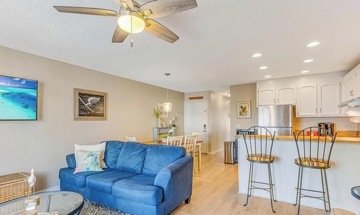 Una sala de estar con un sofá azul, ventilador de techo, televisor y una zona de cocina con taburetes de bar.