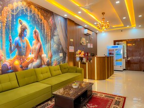 Un vestíbulo de hotel con un sofá verde, un mostrador de recepción, un refrigerador y un mural de deidades.