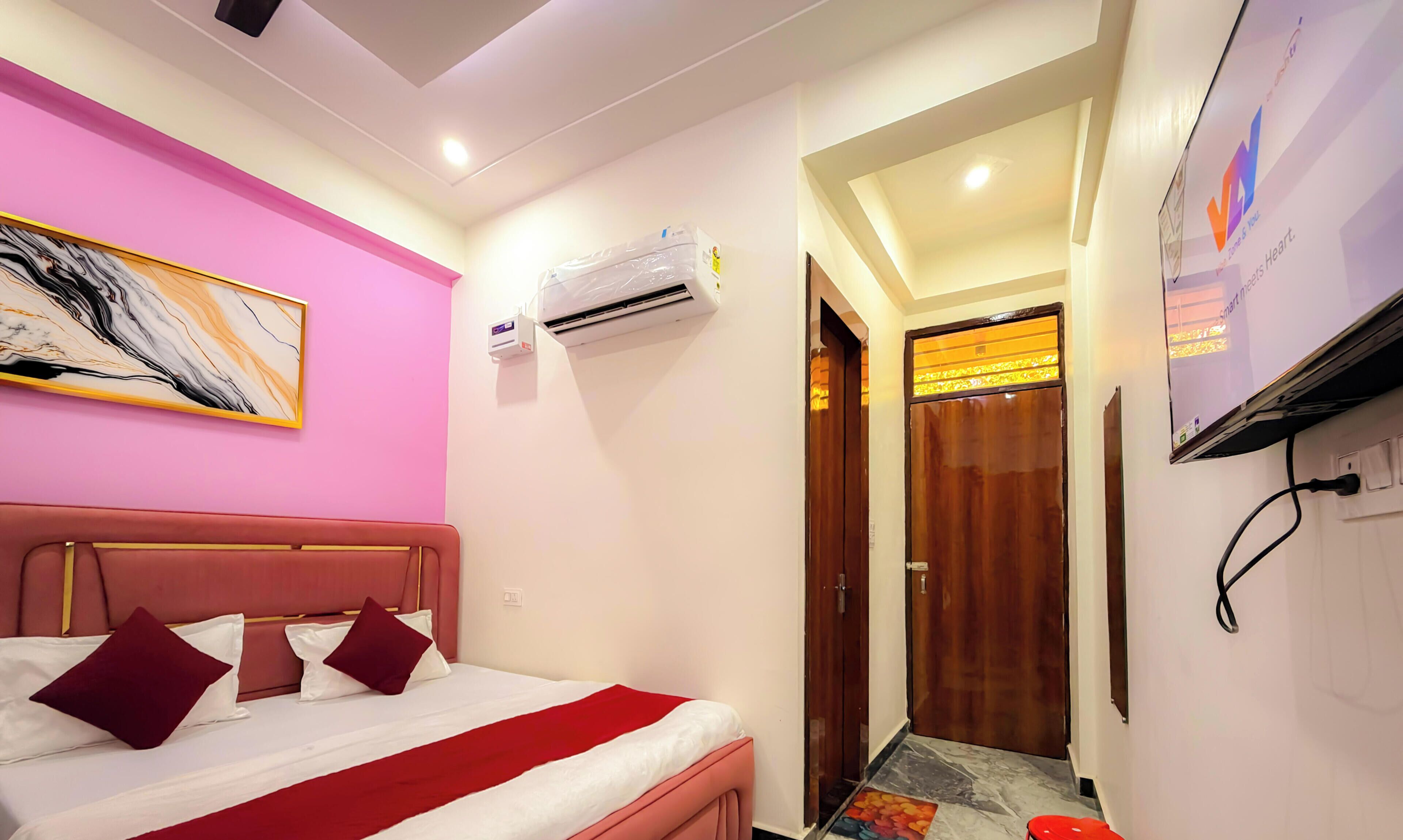 A bedroom with a bed, a painting, an air conditioning unit, a wooden door, and a TV mounted on the wall.