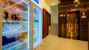 Un hall de hotel con un refrigerador amplio e iluminado lleno de bebidas, un mostrador de recepción de mármol y un ascensor.