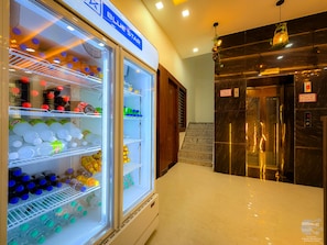 A hotel lobby with a large, well-lit refrigerator stocked with beverages, a marble reception desk, and an elevator. - Radha Palace (Vrindavan)