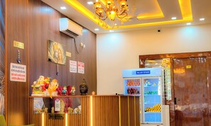 A hotel reception area with a wooden reception desk, a refrigerator stocked with beverages, and a ceiling with a chandelier. - Radha Palace (Vrindavan)