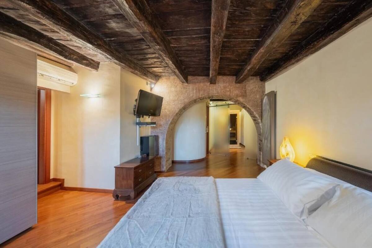 Ein Schlafzimmer mit Holzbalkendecke, einem Fernseher auf einem Schrank, einem Bett mit weißer Bettwäsche und einem Steingewölbe, das in einen anderen Raum führt.