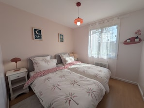 Ein Schlafzimmer mit zwei Betten, einem Nachttisch, einer Lampe, einem Fenster mit Vorhängen und Wandbildern.