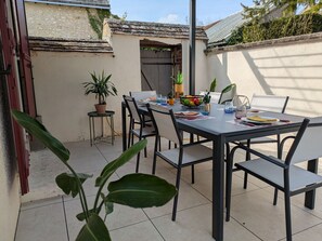 Um pátio ao ar livre com mesa posta para refeições, rodeado por cadeiras e um vaso de planta.