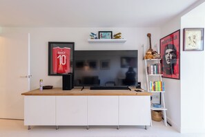 Ein modernes Wohnzimmer mit einem großen Flachbildfernseher, einem Holztisch, einem weißen Schrank und einem Bücherregal mit verschiedenen Gegenständen.