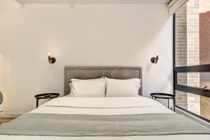 A modern hotel room with a large bed, two bedside tables, a window with a view of a brick building, and a textured throw blanket. - w Amazing Cozy Loft in the Heart of Bogot (Bogotá)