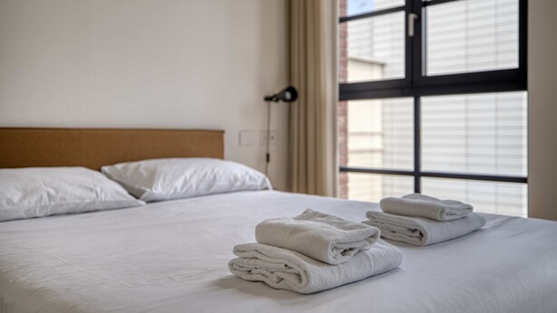 Een hotelkamer met een net opgemaakt bed, een raam met zonwering en een wandlamp.