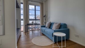 Een moderne woonkamer met een blauwe bank, een klein rond tafeltje, een eethoek met stoelen en een groot raam met gordijnen.