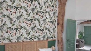 Quarto com cama, cabeceira de vime, porta verde e parede com padrão de folhagem tropical.