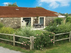 Une maison en pierre avec un toit en tuiles, une clôture en bois et un jardin où poussent des fleurs roses.
