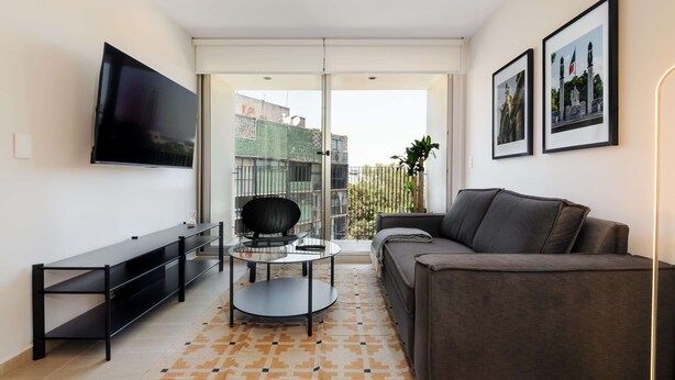 A modern living room with a flat-screen TV, a sofa, a coffee table, and a balcony with a view of buildings. - w Deluxe 2BR w Private Terrace in Hipodromo (Mexico City)