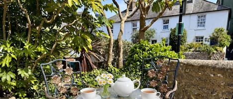 A round metal table with a white teapot, two cups of tea, a vase with daisies, and a plate with scones and jam.
