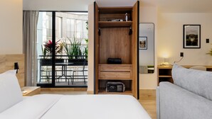 Habitación de hotel con cama, sofá gris, armario de madera, escritorio con lámpara y balcón con plantas.