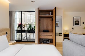 Ein Hotelzimmer mit einem Bett, einem grauen Sofa, einem Holzschrank, einem Schreibtisch mit Lampe und einem Balkon mit Pflanzen.