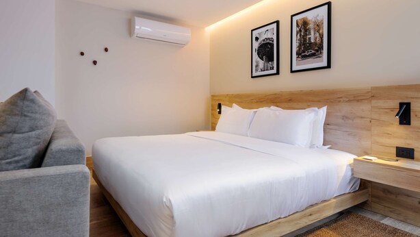 Una habitación de hotel moderna con un cabecero de madera, una cama grande con ropa blanca, un sillón gris y dos cuadros en la pared.