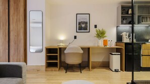 Una habitación moderna con un escritorio de madera, una silla, un pequeño cubo de basura y una zona de cocina con refrigerador y armarios.