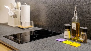 Una cocina con tope de inducción negro, un recipiente blanco para utensilios, un rollo de toallas de papel, botellas de aceite de oliva y sal, y algunos paquetes de especias.