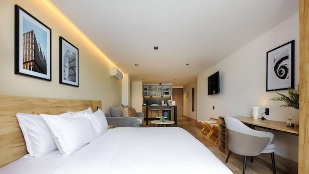 Ein Hotelzimmer mit Bett, Schreibtisch, Stuhl, Fernseher und einer kleinen Küche im Hintergrund.