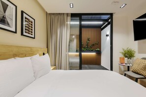 Ein Hotelzimmer mit hölzernem Kopfteil, einem Bett mit weißer Bettwäsche, einer Schiebetür zu einem Balkon mit Pflanzen und einem wandmontierten Flachbildfernseher.